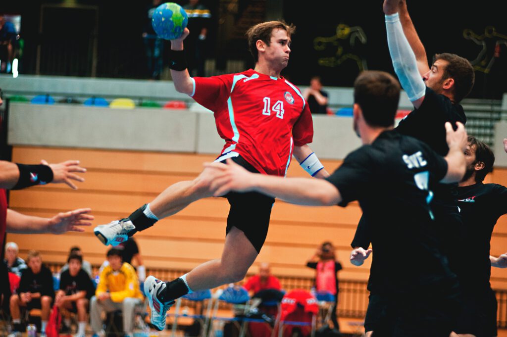 Ein Mann springt in der Luft während eines Handballspiels in einem Sporthaus mit einigen Zuschauern im Hintergrund.